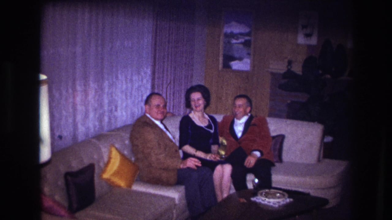 In 1967 Ottawa home-movie footage, three adults sit together on a beige sofa with curtains, pillows, wall art, and a coffee table nearby.