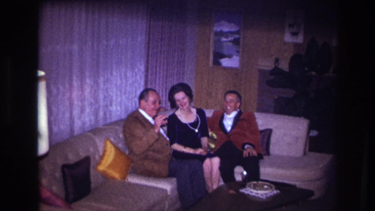 In the Ottawa living room scene, the man in a brown jacket gestures toward the others while a woman in black and a man in red sit beside him.