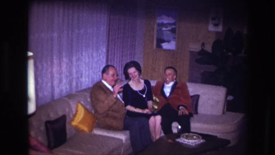 In the Ottawa living room scene, the man in a brown jacket gestures toward the others while a woman in black and a man in red sit beside him.