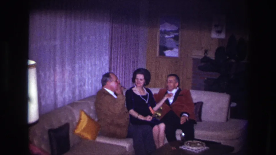 The 1967 Ottawa home-movie frame shows three adults on a sofa, with the woman holding a drink and the two men gesturing during conversation.