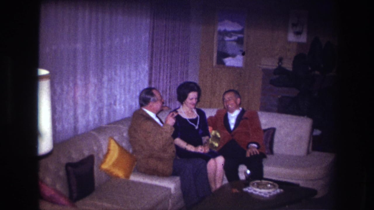In a 1967 Ottawa home-movie frame, two men sit beside a woman on a sofa in a wood-paneled living room.