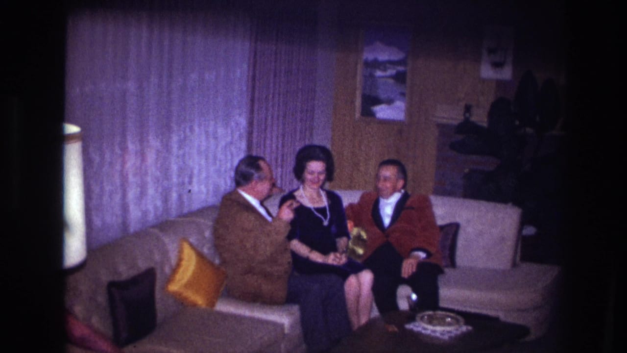 A man in a brown jacket gestures while a woman smiles and another man sits beside her on the sofa.