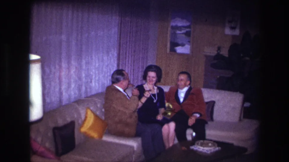 A 1960s home-movie frame shows the seated woman looking down as the two men remain close in the living room.