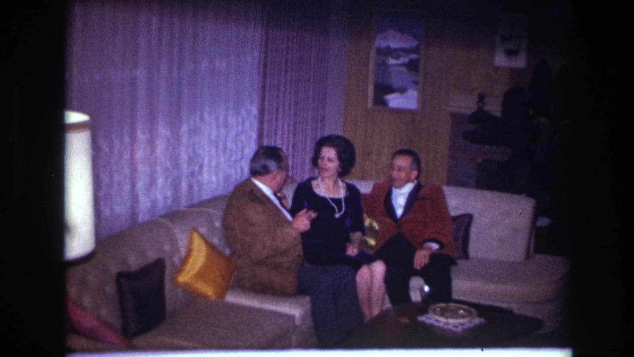 In a 1967 Ottawa home-movie frame, a woman in pearls sits between two men on a curved couch in a wood-paneled living room.