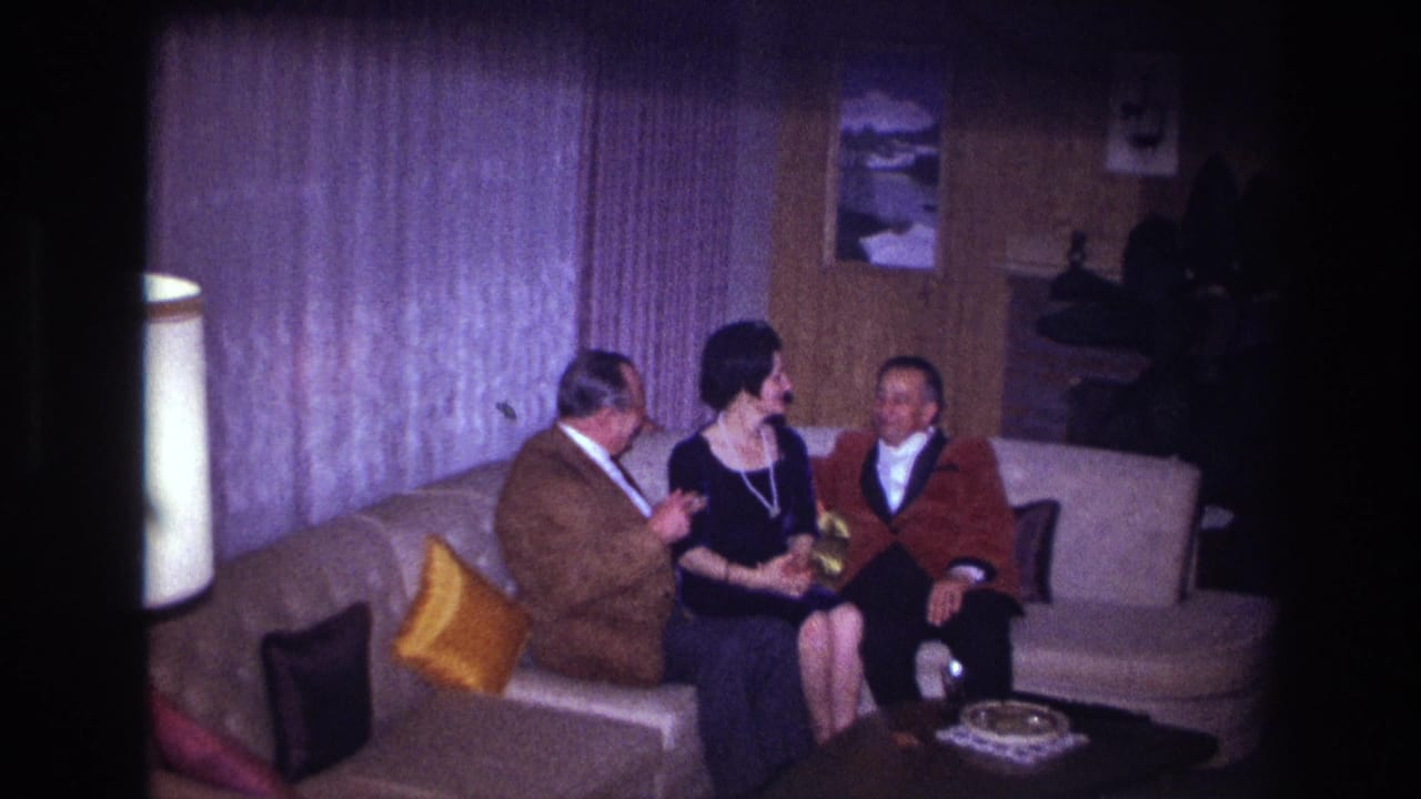 Three adults sit on a light couch in a dim living room with curtains, pillows, and a lamp.