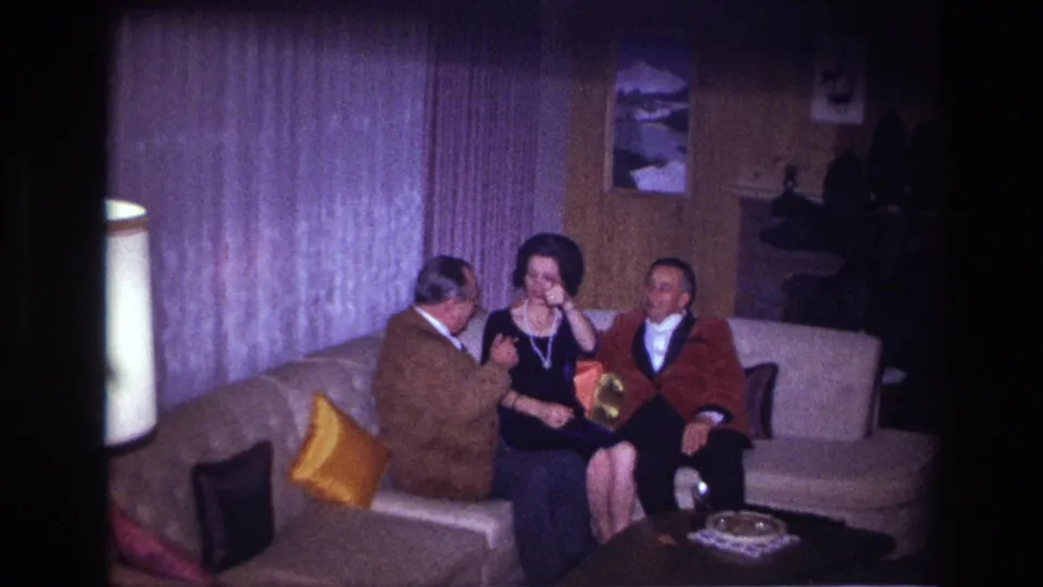 Three adults sit in a private Ottawa living room as the woman in a dark dress raises her hand during the conversation.