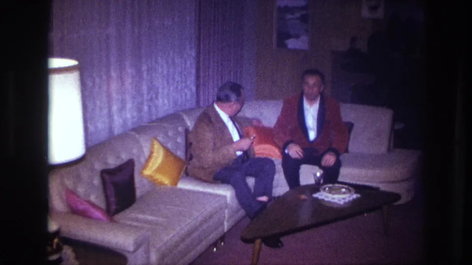 Two men sit on a couch in the 1967 Ottawa home-movie scene, with curtains, pillows, and a coffee table visible nearby.