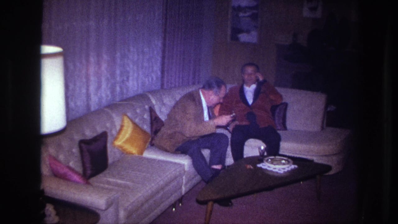 1967 Ottawa home-movie frame of two men conversing on a curved couch in a dim living room with curtains, pillows, lamp, and coffee table.
