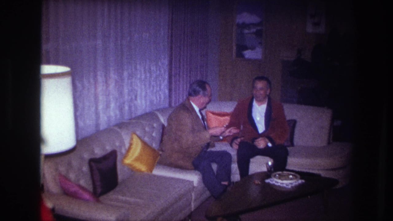 1967 Ottawa interior frame showing two men seated close together on a curved couch during a quiet conversation in a private living room.
