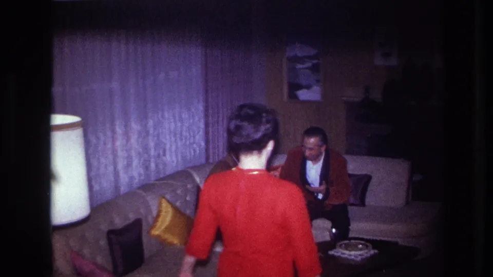 1967 Ottawa home-movie frame of a woman in a red dress entering a dim living room as a seated man remains on the curved couch.
