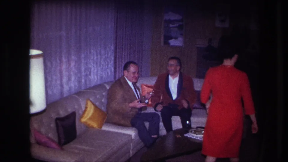 1967 Ottawa living-room frame showing a woman in a red dress approaching two seated men on a curved couch with mid-century decor around them.