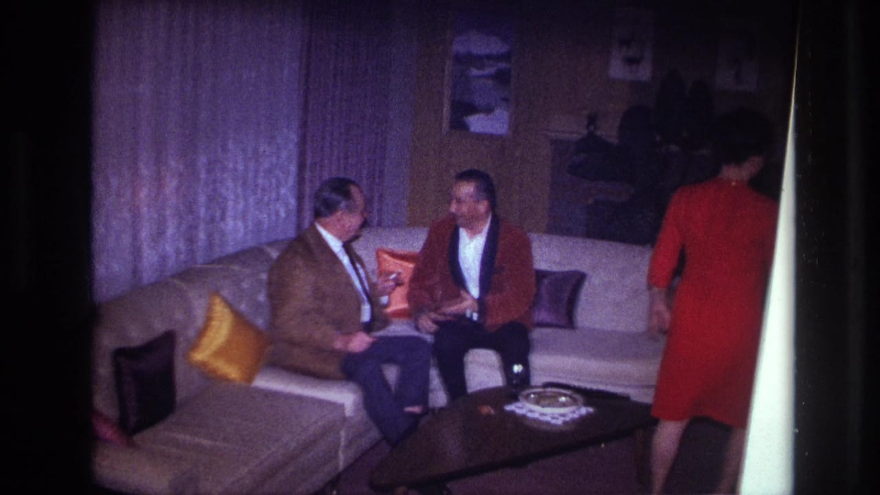 In a 1967 Ottawa home interior, two formally dressed men talk on a beige sectional as a woman in a red dress passes by.