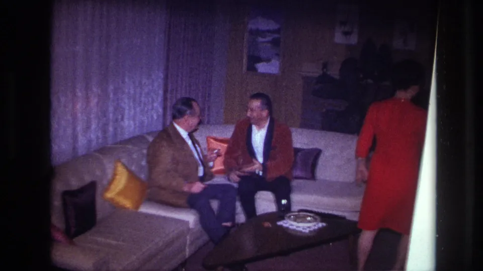 In a 1967 Ottawa home interior, two formally dressed men talk on a beige sectional as a woman in a red dress passes by.