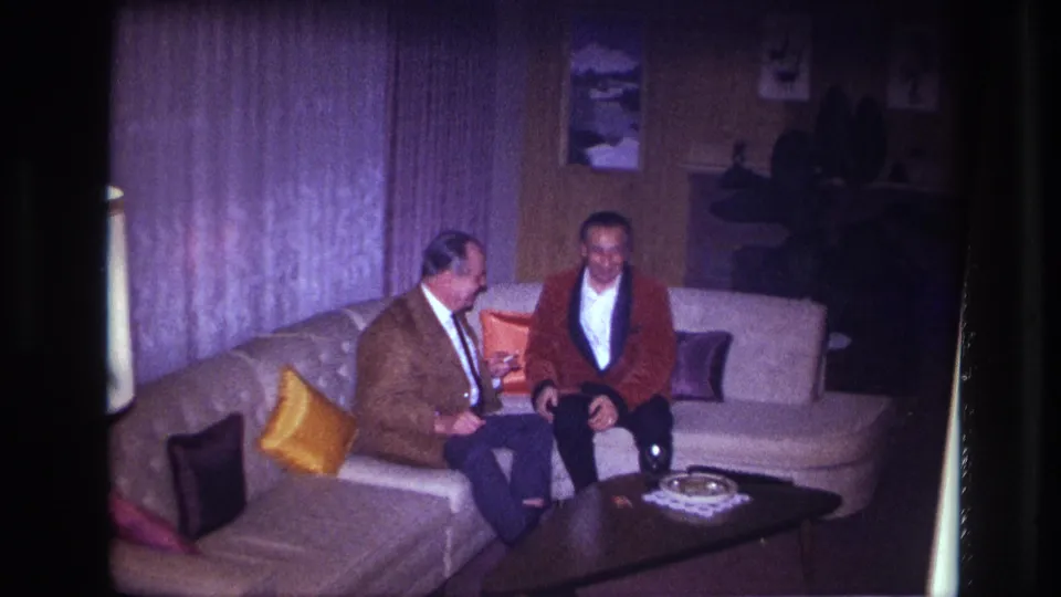 Two men in formal clothing sit on a beige sectional in a dim 1967 Ottawa living room with pillows, curtains, and a coffee table.