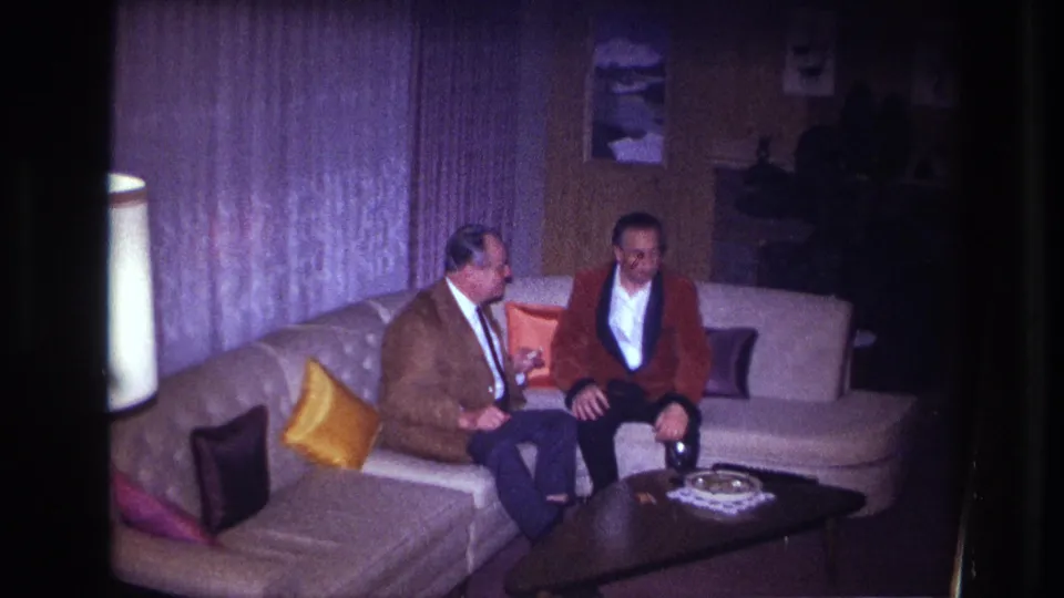 The seated men continue their conversation in the Ottawa living room, framed by curtains, a lamp, pillows, and a dark coffee table.