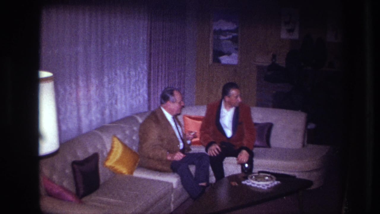 In a 1967 Ottawa home-movie frame, two formally dressed men sit on a sectional couch beside bright pillows and a dark coffee table.
