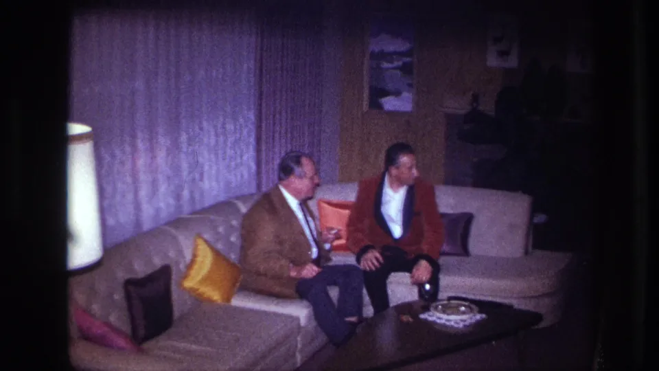 In a 1967 Ottawa home-movie frame, two formally dressed men sit on a sectional couch beside bright pillows and a dark coffee table.