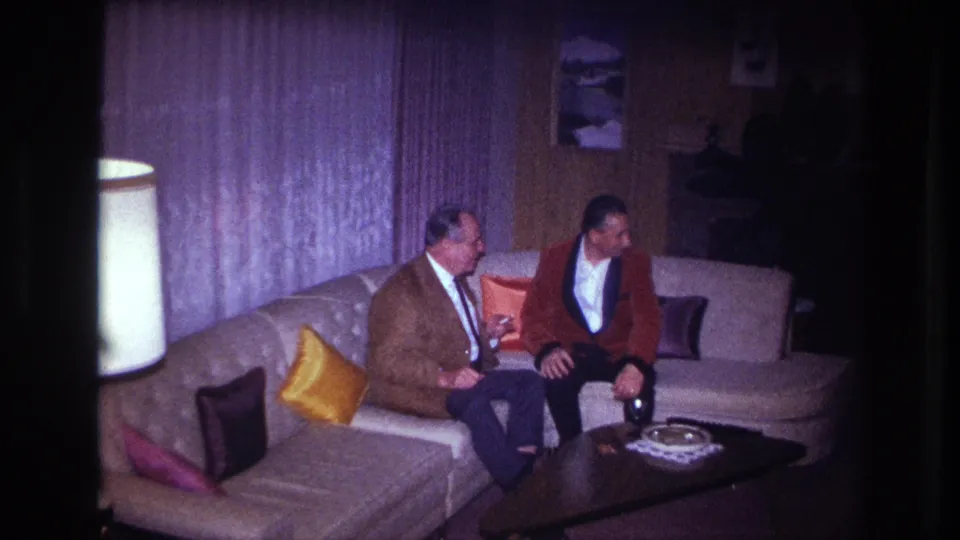 Two men in formal jackets sit in conversation on a couch in a dim 1967 Ottawa living room, looking toward the right side of the room.