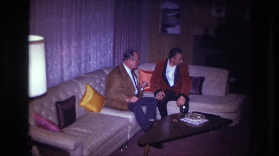 A woman in a red dress appears at the right edge while two men sit on a couch in the dim Ottawa living-room scene.