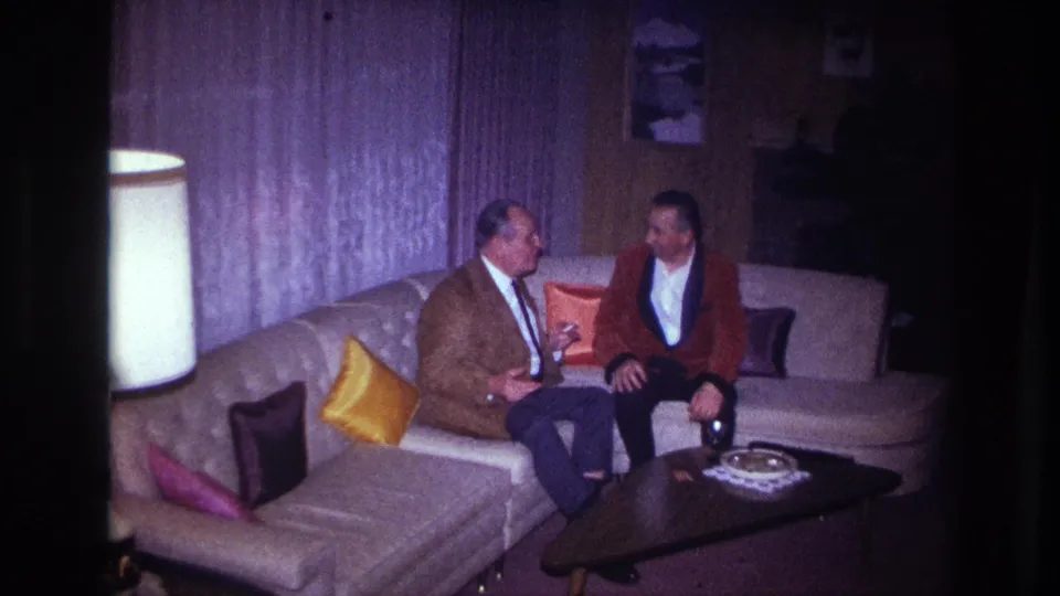 The woman in red stands partly in frame near two seated men on a sectional couch in the 1967 Ottawa home-movie interior.