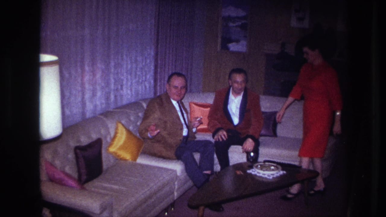 1967 Ottawa home-movie frame of two suited men on a light sofa as a woman in a red dress stands near a coffee table.