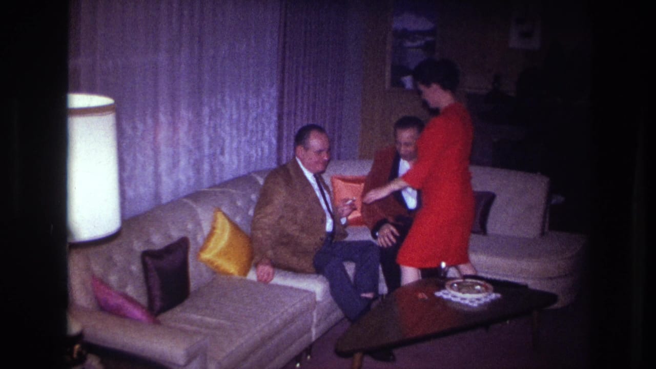 A woman in a red dress stands close to two men seated on a living room sofa.