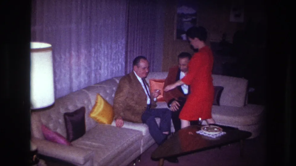 1967 Ottawa interior frame with a woman gesturing near two seated men on a sofa beside pillows and a coffee table.
