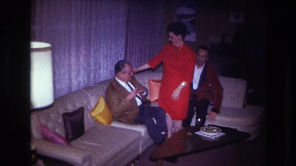1967 Ottawa home-movie frame showing a woman in a red dress moving between two men seated in a living room.