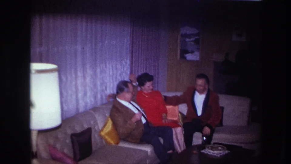1967 Ottawa home-movie frame of a woman seated between two men on a sectional sofa in a private living room.