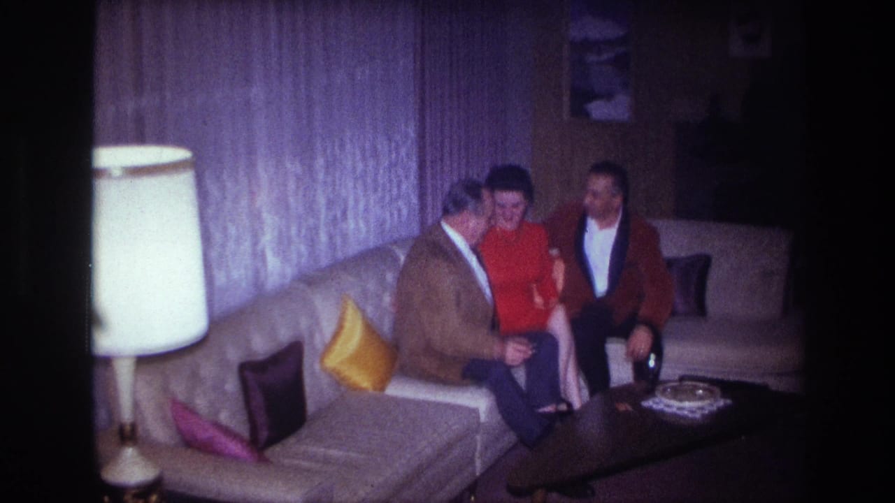 Three adults sit close together on a sectional couch in a 1967 Ottawa living room with curtains and a lamp visible.