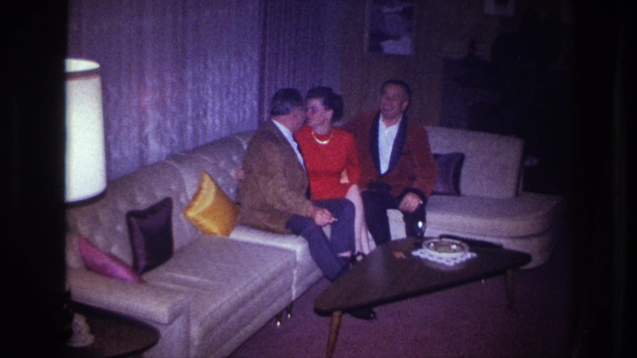 1967 Ottawa home-movie frame showing three adults seated on a mid-century couch, with a woman in red between two men.
