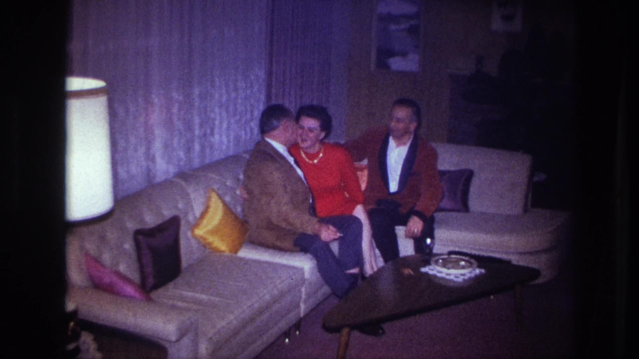 A man leans toward a woman in red while another man sits beside her on the couch.