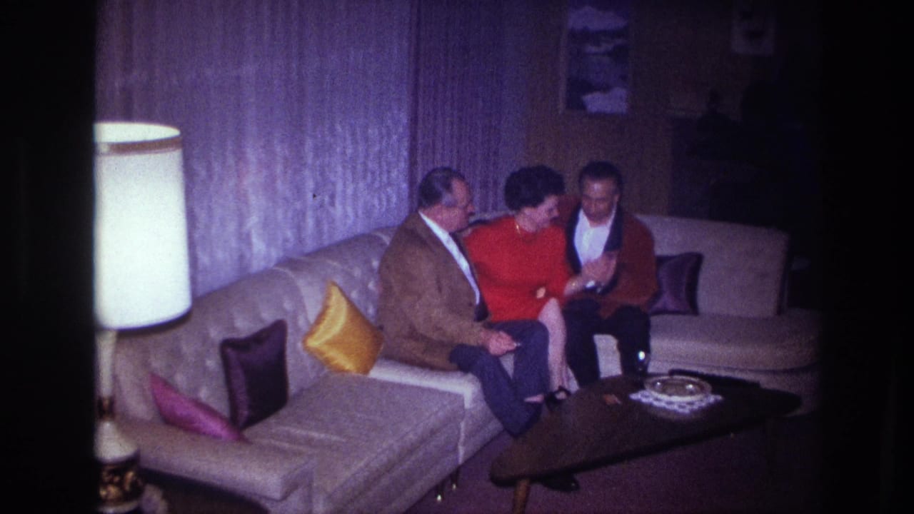 The three adults appear to laugh together on the couch, framed by a lamp, curtains, pillows, and a low coffee table.