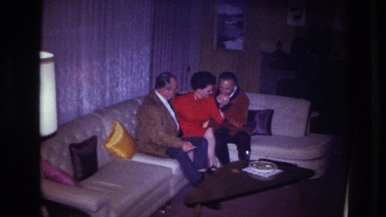 1967 Ottawa home-movie frame showing two men and a woman seated close together on a pale sectional couch in a dim living room.