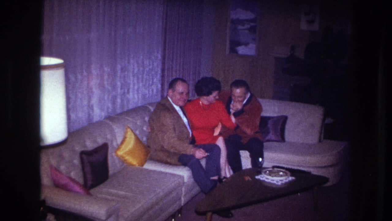 Two men and a woman sit shoulder to shoulder on a light couch, with a coffee table, lamp, curtains, and pillows visible.