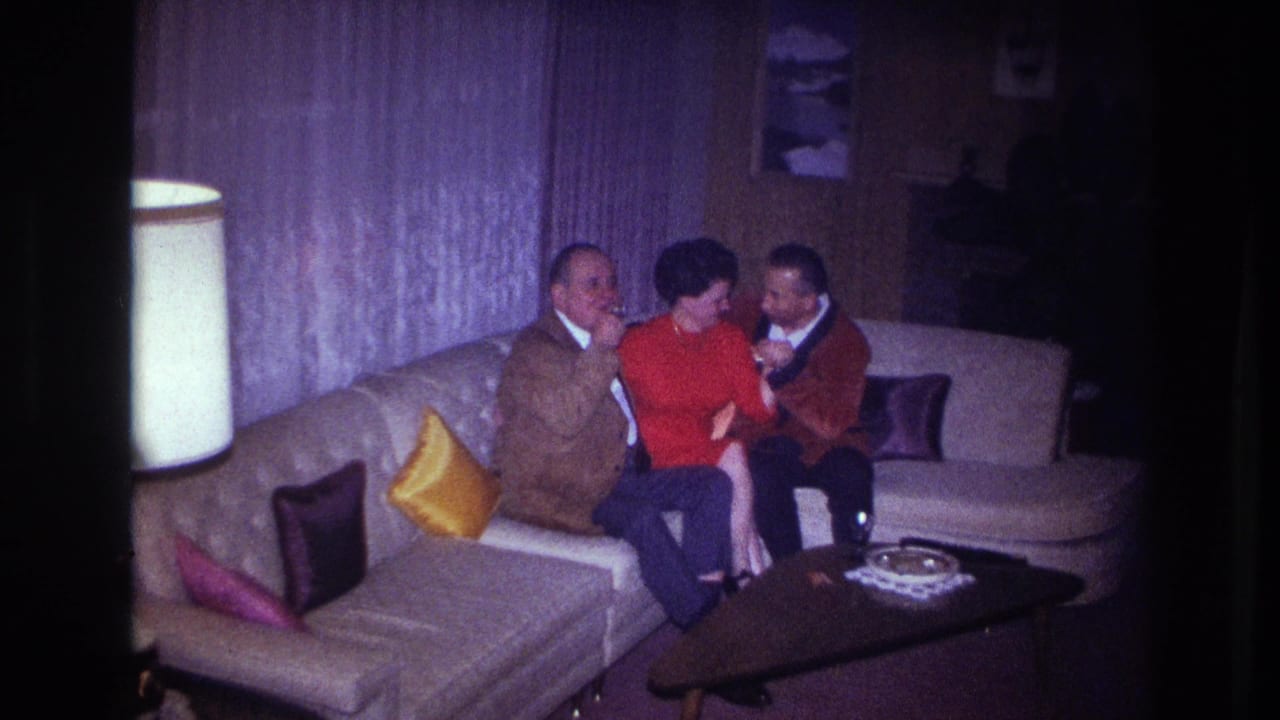 Three adults in 1960s home-movie footage sit on a sectional couch, with the woman in red between two men in jackets.
