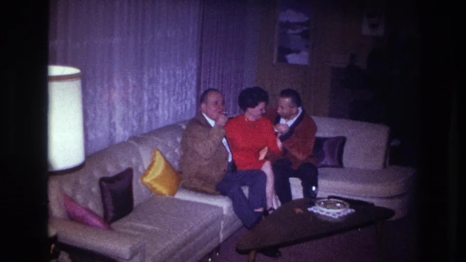 Three adults in 1960s home-movie footage sit on a sectional couch, with the woman in red between two men in jackets.