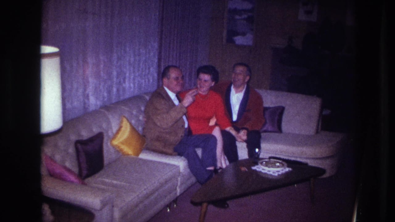 In a dim Ottawa living room, a seated man gestures toward the woman in red as another man sits nearby on the sectional couch.