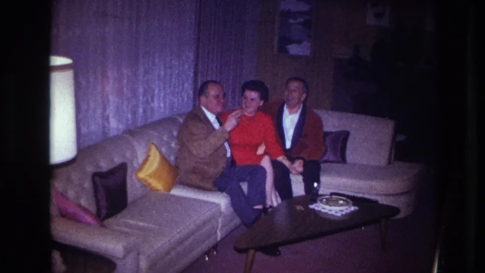 In a dim Ottawa living room, a seated man gestures toward the woman in red as another man sits nearby on the sectional couch.