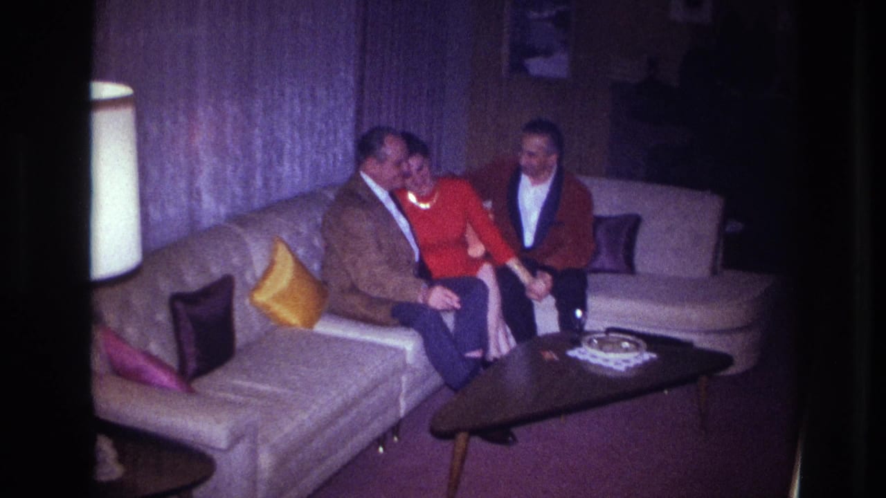 1967 Ottawa home-movie frame showing a woman in red seated between two men on a sectional sofa in a private living room.