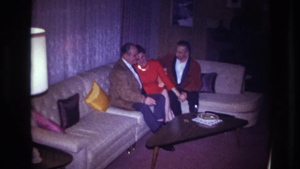 1967 Ottawa home-movie frame showing a woman in red seated between two men on a sectional sofa in a private living room.