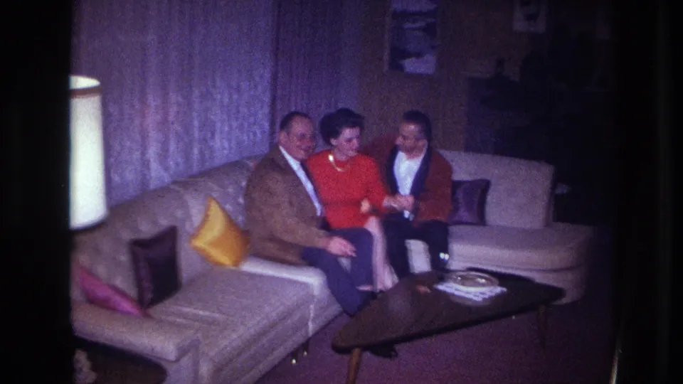 1967 Ottawa home-movie frame showing three adults laughing together on a sofa, with mid-century furnishings visible around them.