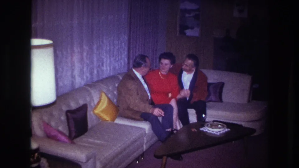 1967 Ottawa home-movie frame of three adults smiling on a sofa in a private living room with curtains, pillows, lamp, and coffee table.