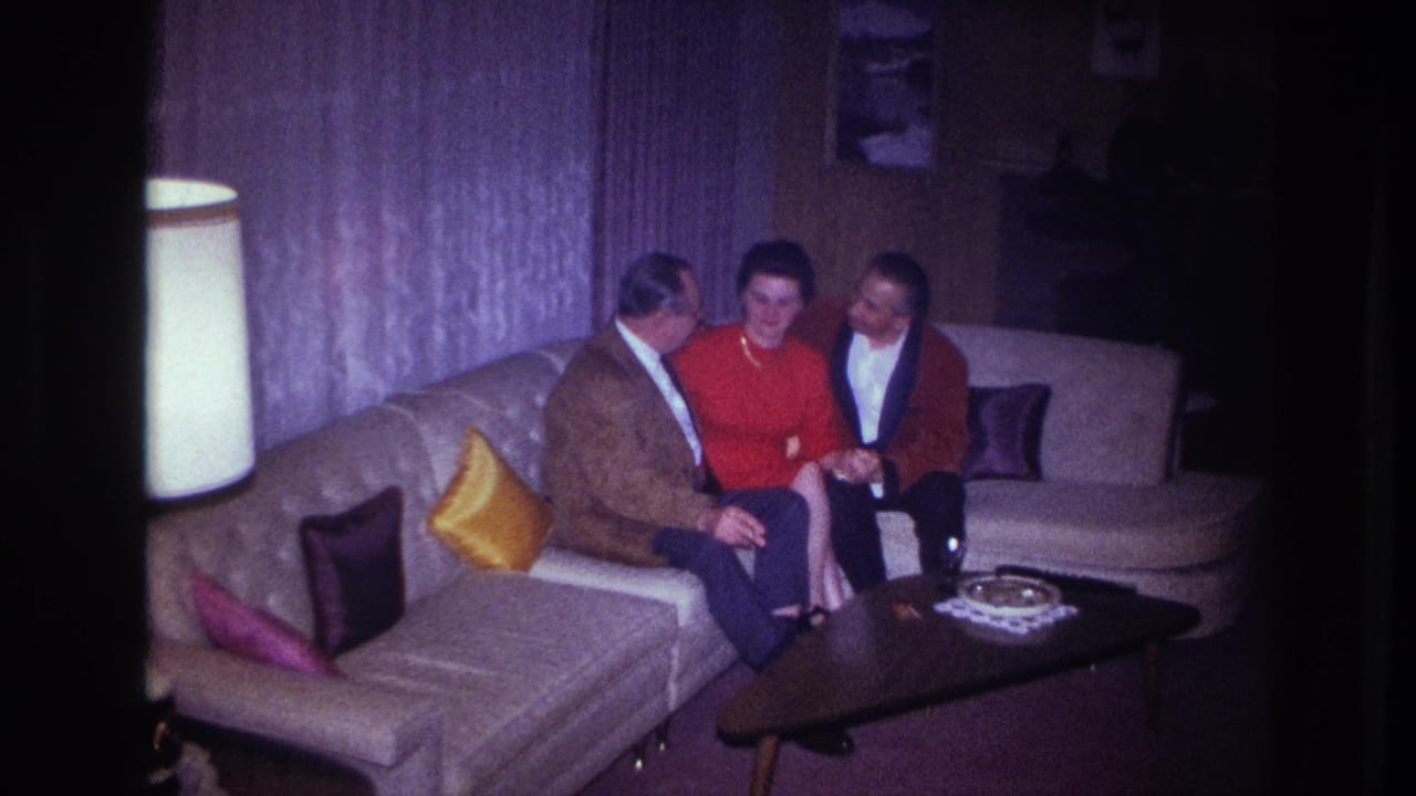 Three adults in an Ottawa apartment interior in 1967 sit together on a light sectional sofa beside a lamp and coffee table.