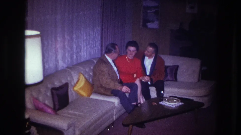 Three adults in an Ottawa apartment interior in 1967 sit together on a light sectional sofa beside a lamp and coffee table.