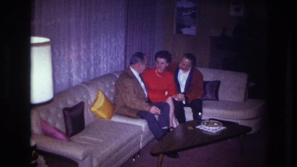 In a 1967 Ottawa home-movie frame, three adults sit on a sectional sofa near a lamp, curtains, pillows, and a low table.
