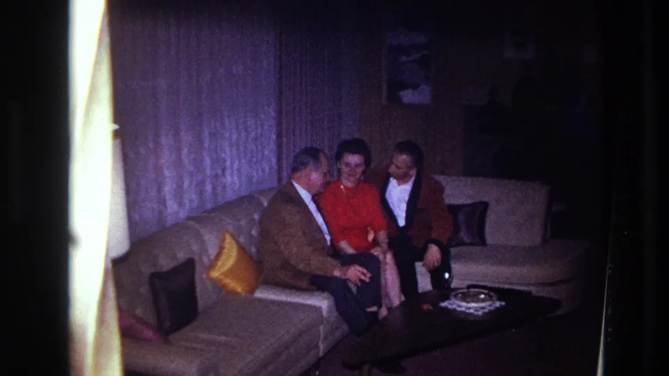 Three adults appear in conversation on a sofa in the Ottawa apartment, with film-edge flare and low interior light visible.
