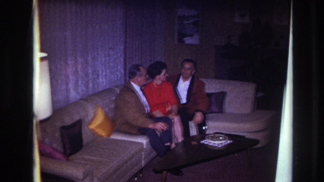1967 Ottawa home-movie frame showing a woman in red seated between two men on a sectional couch in a private living room.