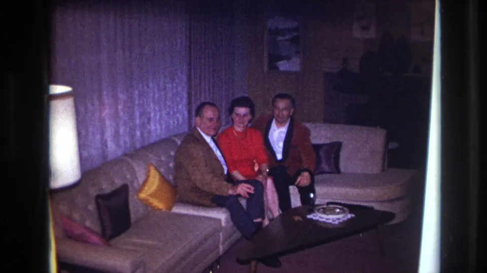 1967 Ottawa home-movie frame of three adults on a sectional couch, with a dark coffee table and glass ashtray in the foreground.