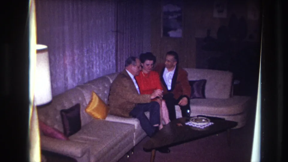 1967 Ottawa home-movie frame showing three adults in conversation on a couch inside a softly lit private living room.